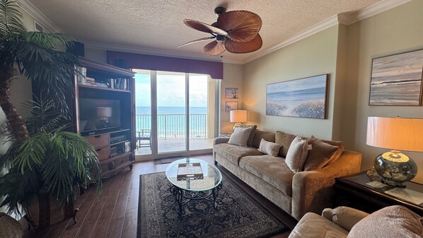 A living room with a sofa, a glass coffee table, a bookshelf, a large window with a view of the ocean, and a ceiling fan.
