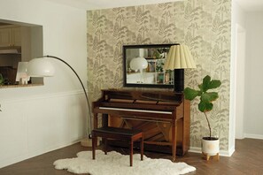 Un piano avec un banc, une lampe et une plante en pot, dans une pièce dont le papier peint est orné de motifs.