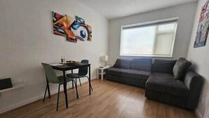 Uma sala de estar com um sofá cinza escuro, uma mesa de jantar com duas cadeiras, uma janela com persianas e um quadro abstrato colorido na parede.