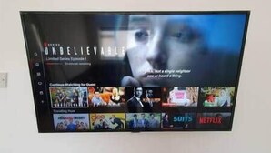 Televisão de tela plana presa na parede exibindo a interface de um serviço de streaming com diversas opções de conteúdo.