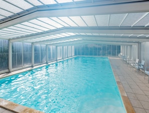 Uma piscina interior com paredes de vidro transparente, chão ladrilhado e uma fileira de cadeiras brancas ao longo da lateral.