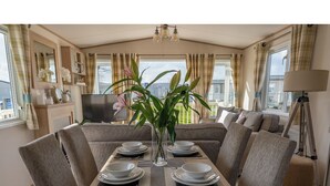A dining area with a table set for four, surrounded by upholstered chairs, a television, and a large window with curtains. - A luxury Caravan in a suburb location. 
       AJH Holiday Homes, Ty Mawr. (Towyn)