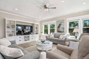 A modern living room with a flat-screen TV, grey sofas, a glass coffee table, and a ceiling fan. - Sea The Pointe: Luxurious Living on Davis Canal (Oak Island)