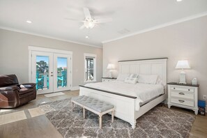 Quarto com cama, mesas de cabeceira, um banco, ventoinha no teto e vista através de uma porta de vidro.