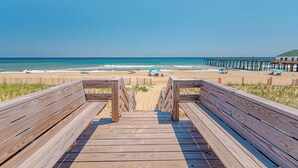 Eine Holzpromenade führt zu einem Strand, im Hintergrund ist ein Steg zu sehen.