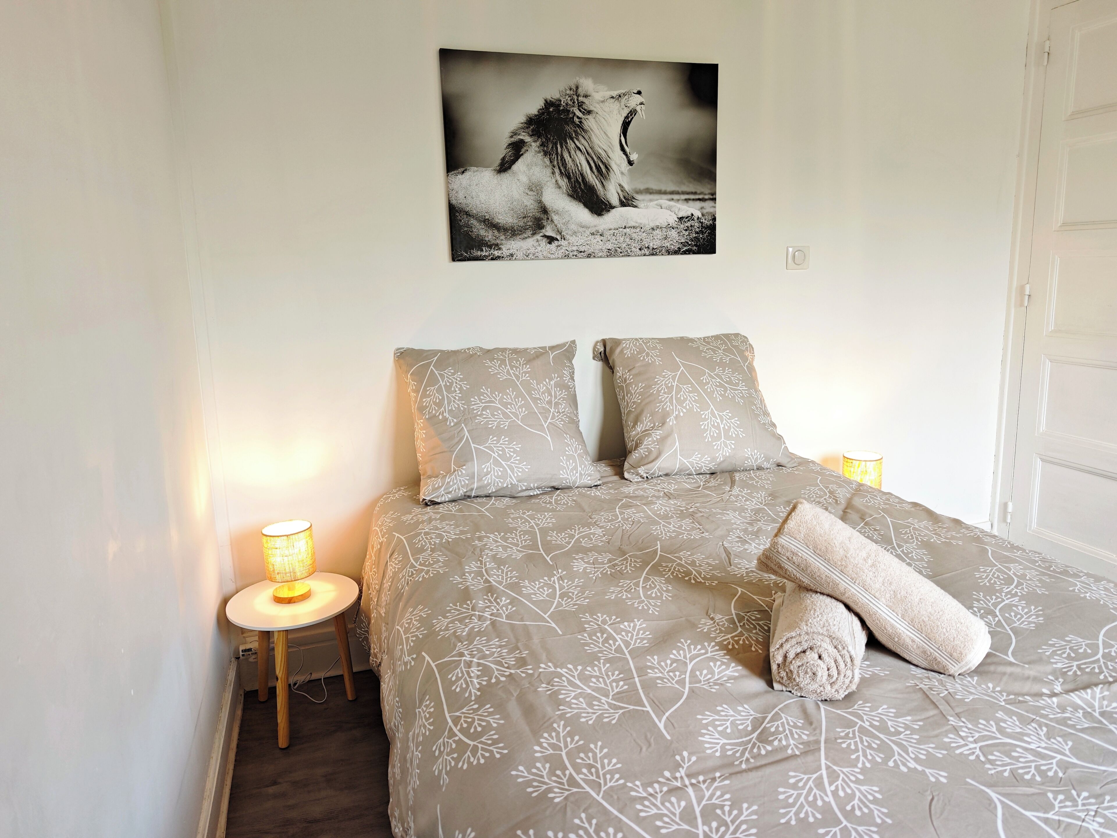 A bedroom with a bed, two pillows, a blanket, a rolled towel, a bedside lamp, and a framed picture on the wall.