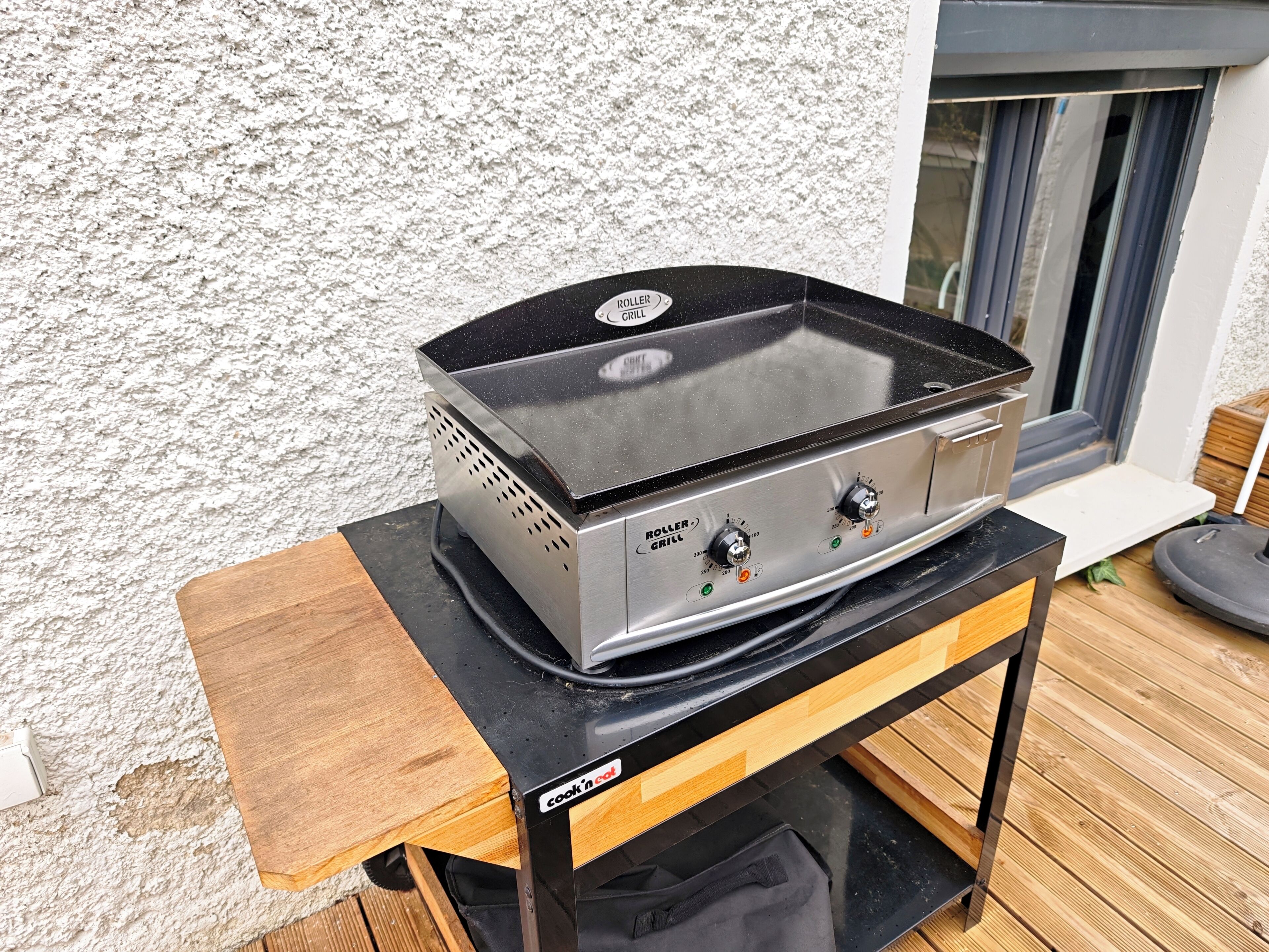 Ein tragbarer Grill auf einem Holztisch, im Hintergrund eine strukturierte Wand und ein Fenster.