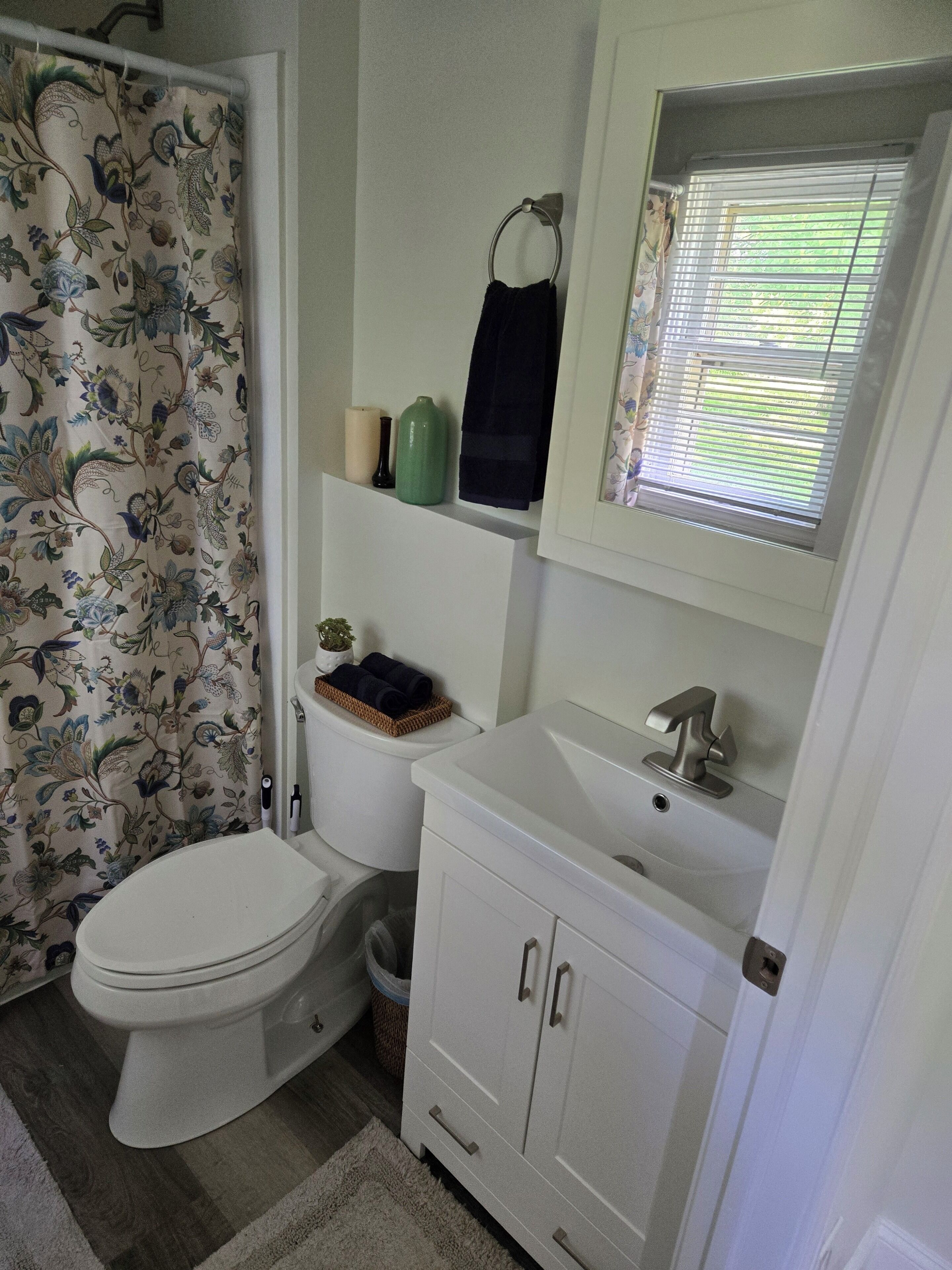 A bathroom with a toilet, a sink, a cabinet, a shower curtain, and a window with blinds.