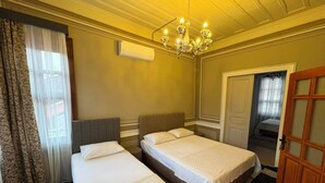 A hotel room with two single beds, a chandelier, an air conditioning unit, a door, and a window with curtains.