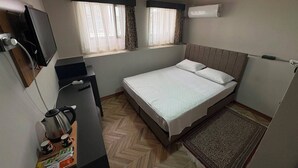 A compact hotel room with a bed, a desk with a kettle and cups, a television, and an air conditioning unit.