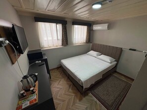 A compact hotel room with a bed, a desk with a kettle and cups, a television, and an air conditioning unit.