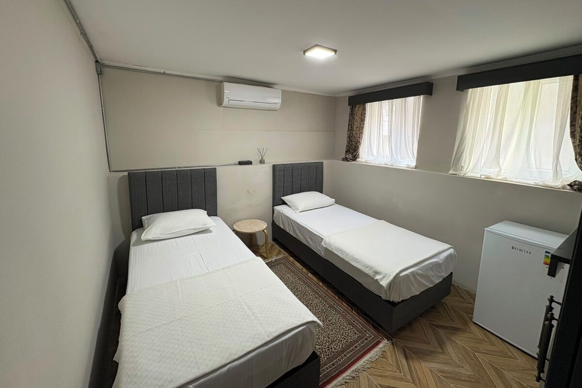 A hotel room with two single beds, a small table, a refrigerator, and a window with curtains.