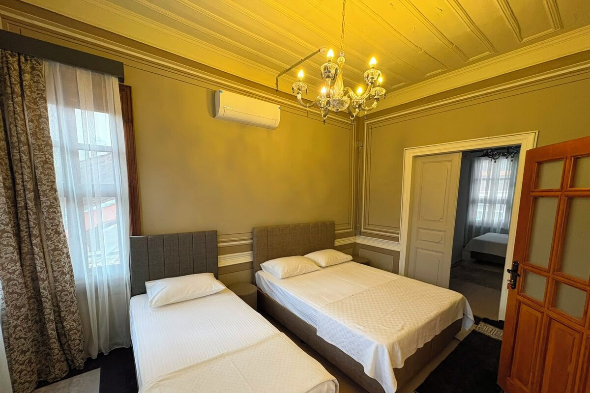 A hotel room with two single beds, a chandelier, an air conditioning unit, a door, and a window with curtains.