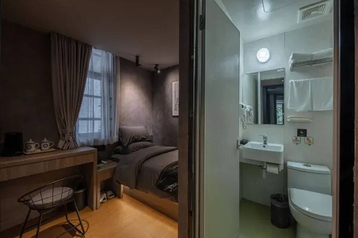 Una habitación funcional con cama, escritorio y silla, ventana con cortinas y baño con lavamanos e inodoro.