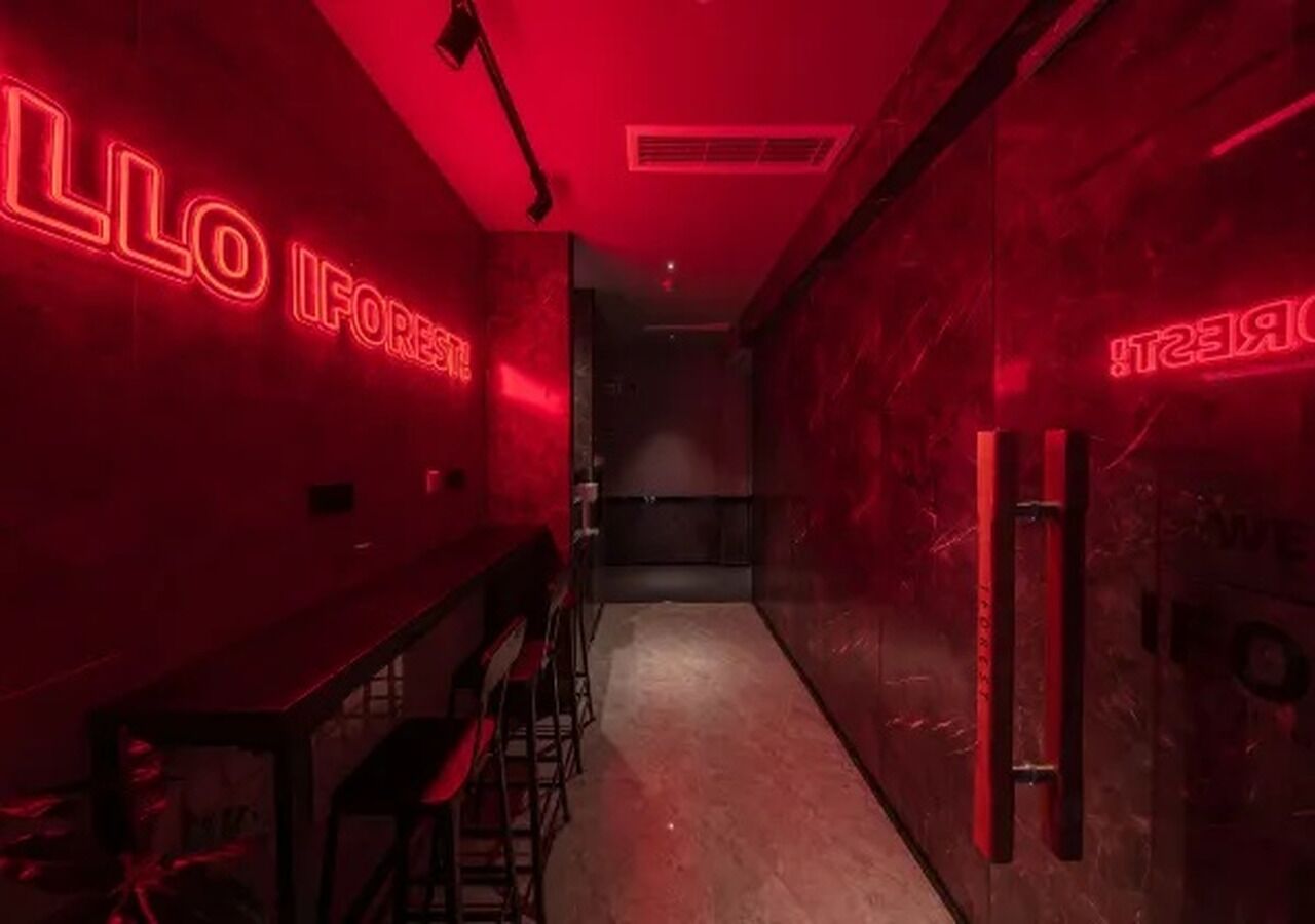 Un corridoio illuminato di rosso con un'area bar, caratterizzata da insegne al neon, un bancone e sgabelli da bar.