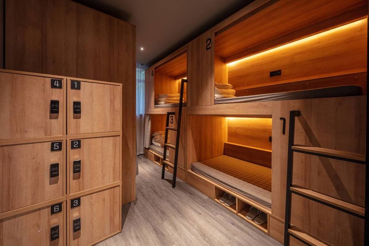 Filas de literas de madera con números que señalan las habitaciones, una escalera y un muro de madera con luz cálida.