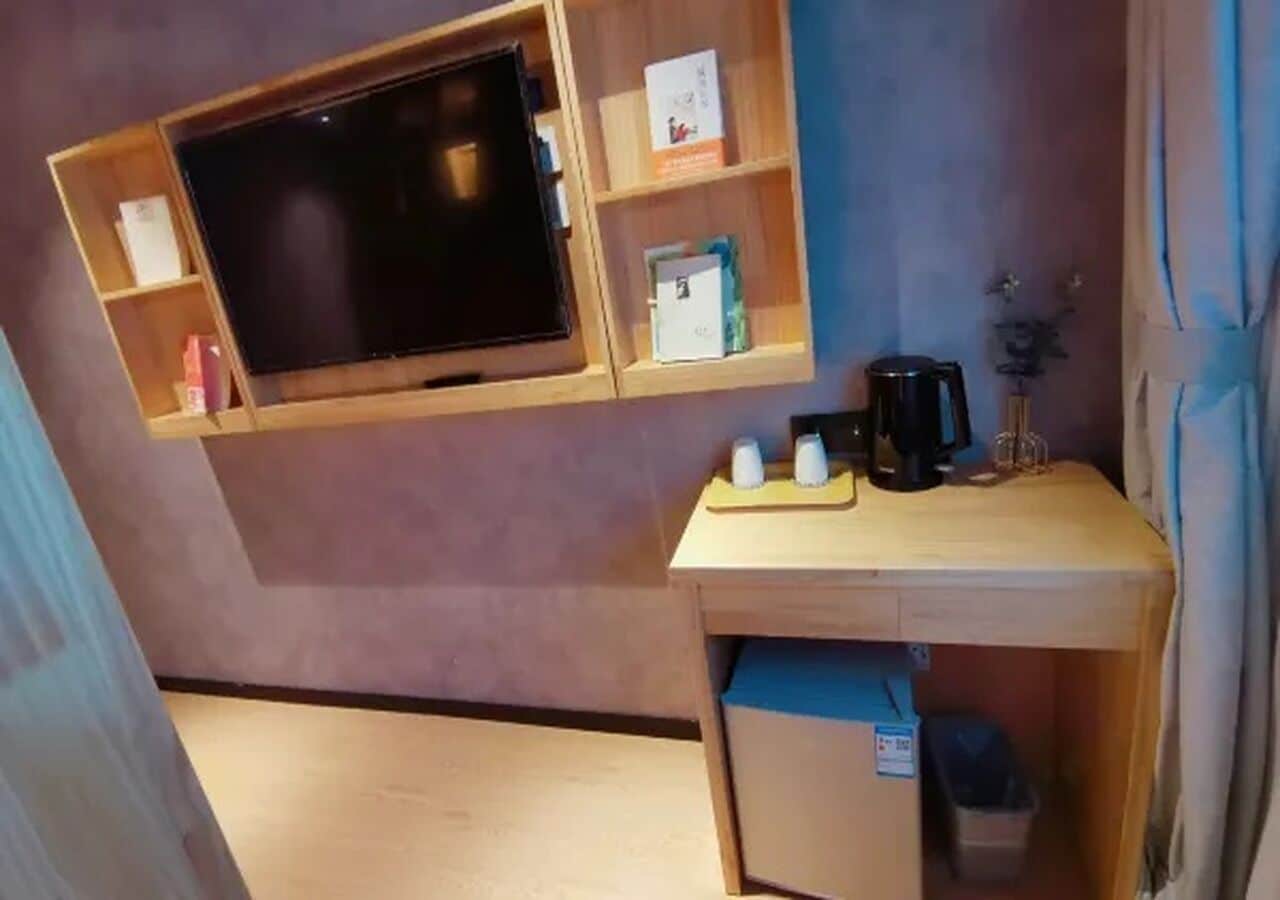A compact room with a mounted television, a small wooden desk, and a trash bin.