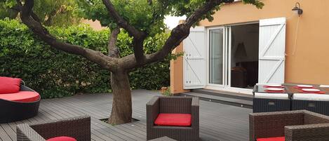An outdoor patio with wicker furniture and red cushions, a tree, and a building with a white door and shutters.