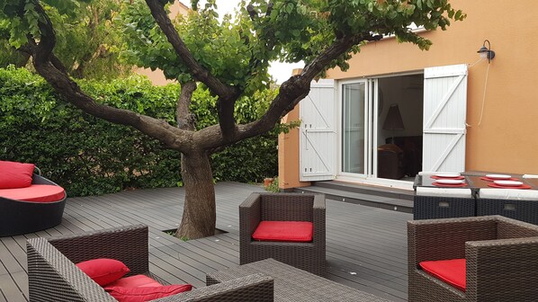 An outdoor patio with wicker furniture and red cushions, a tree, and a building with a white door and shutters. - 300m from the sea, 75 m² enclosed garden apartment for 4 people (La Seyne-sur-Mer)