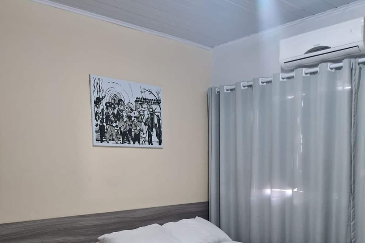 A bedroom with a bed, a wall-mounted air conditioner, a window with curtains, and a framed picture on the wall.