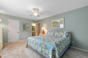 A bedroom with a bed, a ceiling fan, a nightstand, a lamp, a door, a mirror, and a wall with a framed picture. - North Myrtle Beach Cottage 2 Blocks From Beach (North Myrtle Beach)