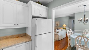 Una cocina con gabinetes blancos, lavavajillas integrado y refrigerador. Hay un comedor con una mesa, sillas, una lámpara y un candelabro.