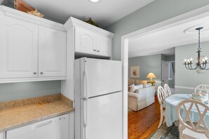 A kitchen with white cabinetry, a built-in dishwasher, and a refrigerator. There is a dining area with a table and chairs, a lamp, and a chandelier. - North Myrtle Beach Cottage 2 Blocks From Beach (North Myrtle Beach)