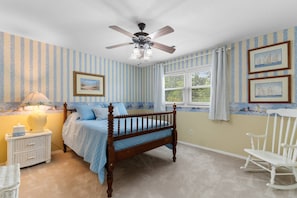 A bedroom with a bed, a nightstand, a lamp, a chair, a ceiling fan, and a window with curtains. - North Myrtle Beach Cottage 2 Blocks From Beach (North Myrtle Beach)
