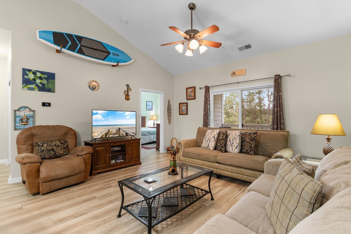 A living room with a sofa, armchair, TV, ceiling fan, and a surfboard decoration.