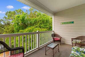 A covered porch with a white railing, two wicker chairs with cushions, a glass-top table, and a sign reading 'MARGARITAVILLE'. - Top-Floor Barefoot retreat | Nature views + Pool (North Myrtle Beach)