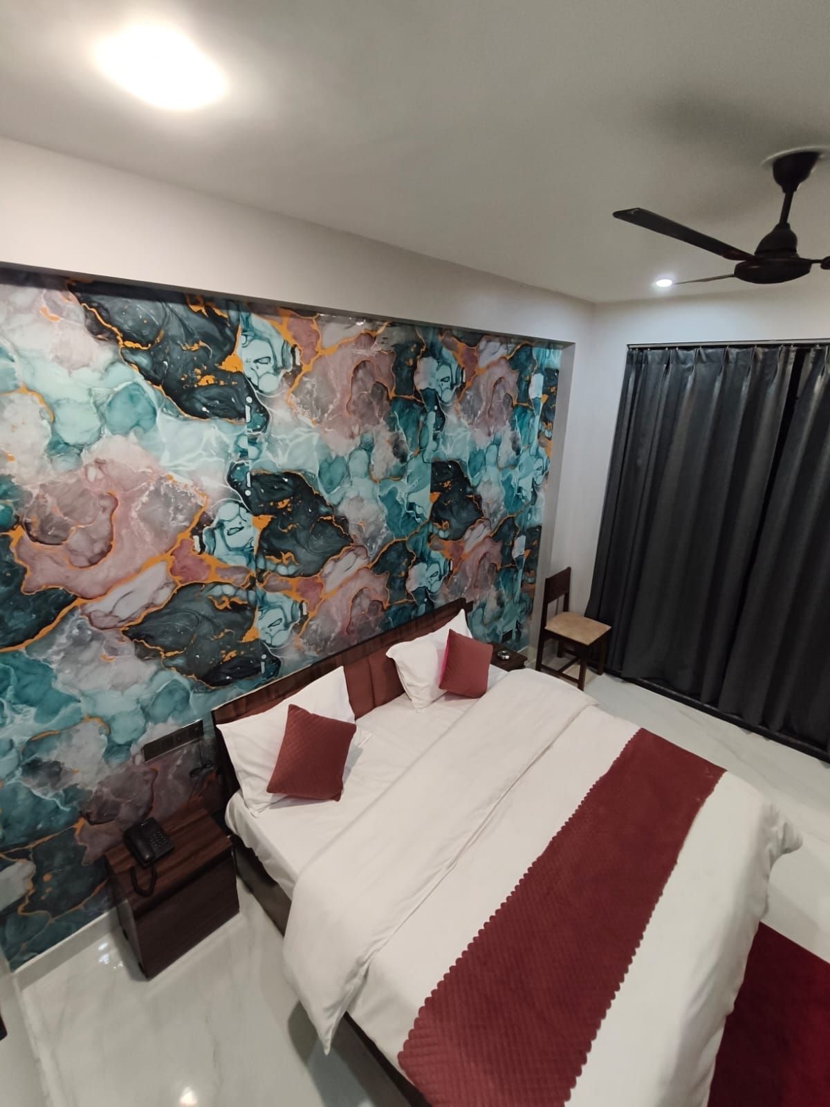 A hotel room with a bed, a nightstand, a chair, a ceiling fan, and a large abstract painting on the wall.