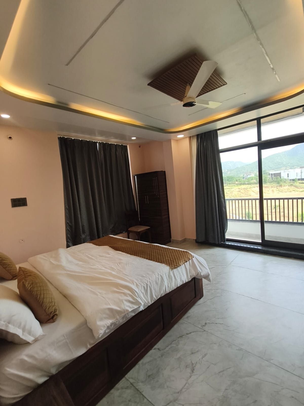 A hotel room with a large bed, a flat-screen TV, a ceiling fan, and a view of a mountainous landscape through the windows.