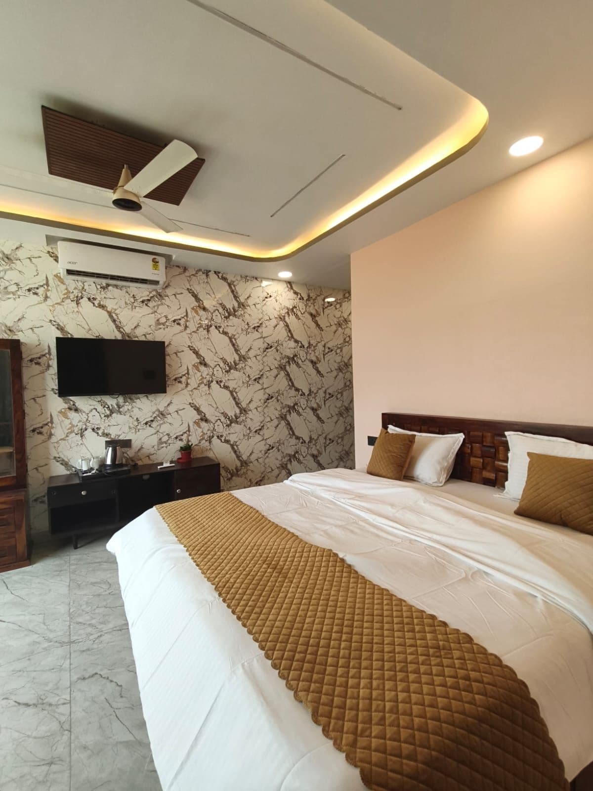 A hotel room with a bed, a flat-screen TV mounted on the wall, a ceiling fan, and an air conditioning unit.
