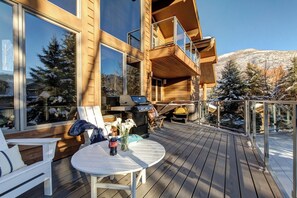 Um deck de madeira com área de estar ao ar livre, churrasqueira e vista para árvores cobertas de neve.
