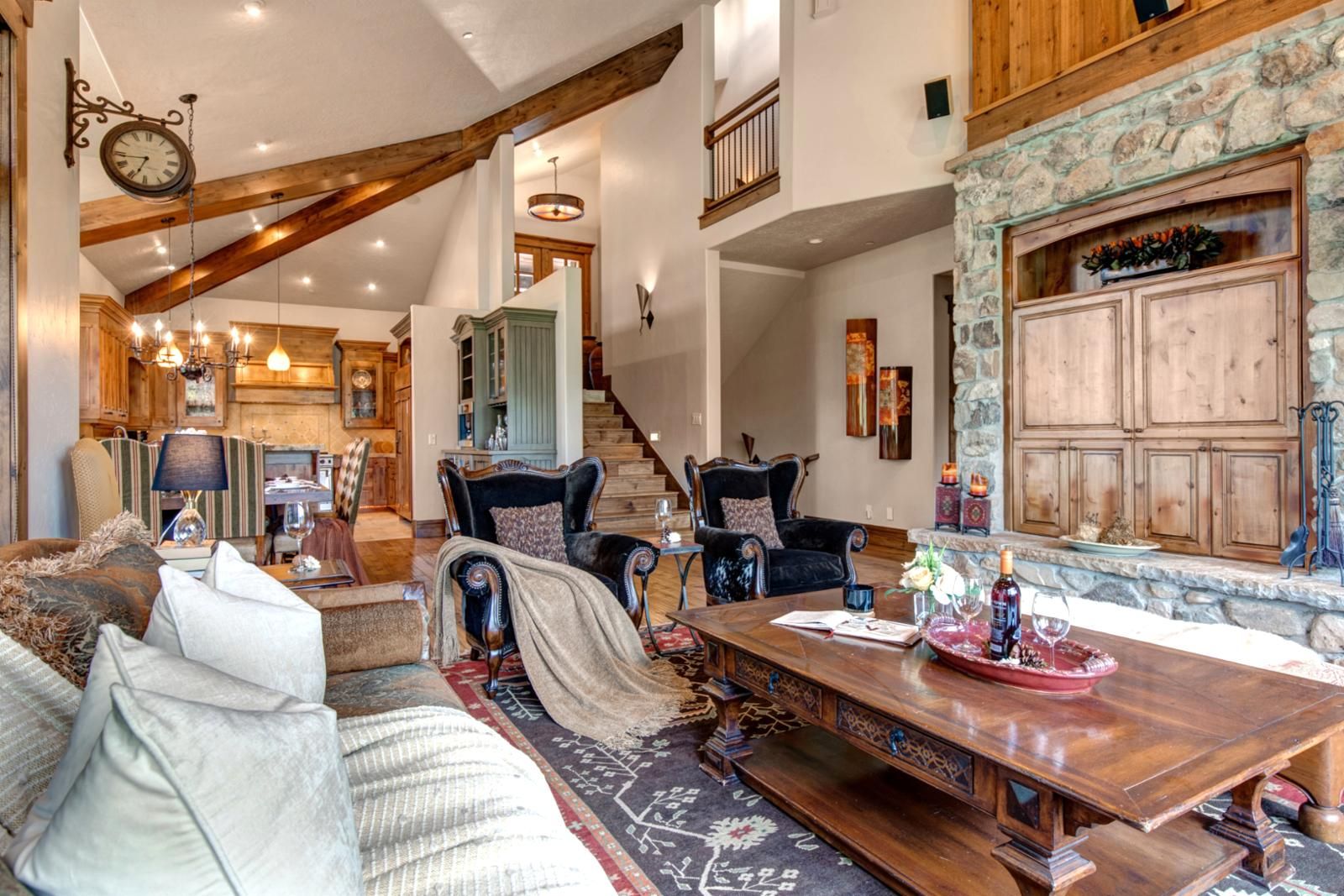 A spacious living area with a high ceiling, wooden beams, a stone fireplace, and a dining table set with wine glasses and a tray.