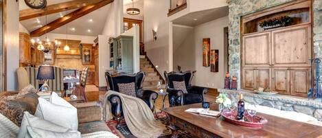 A spacious living area with a high ceiling, wooden beams, a stone fireplace, and a dining table set with wine glasses and a tray.