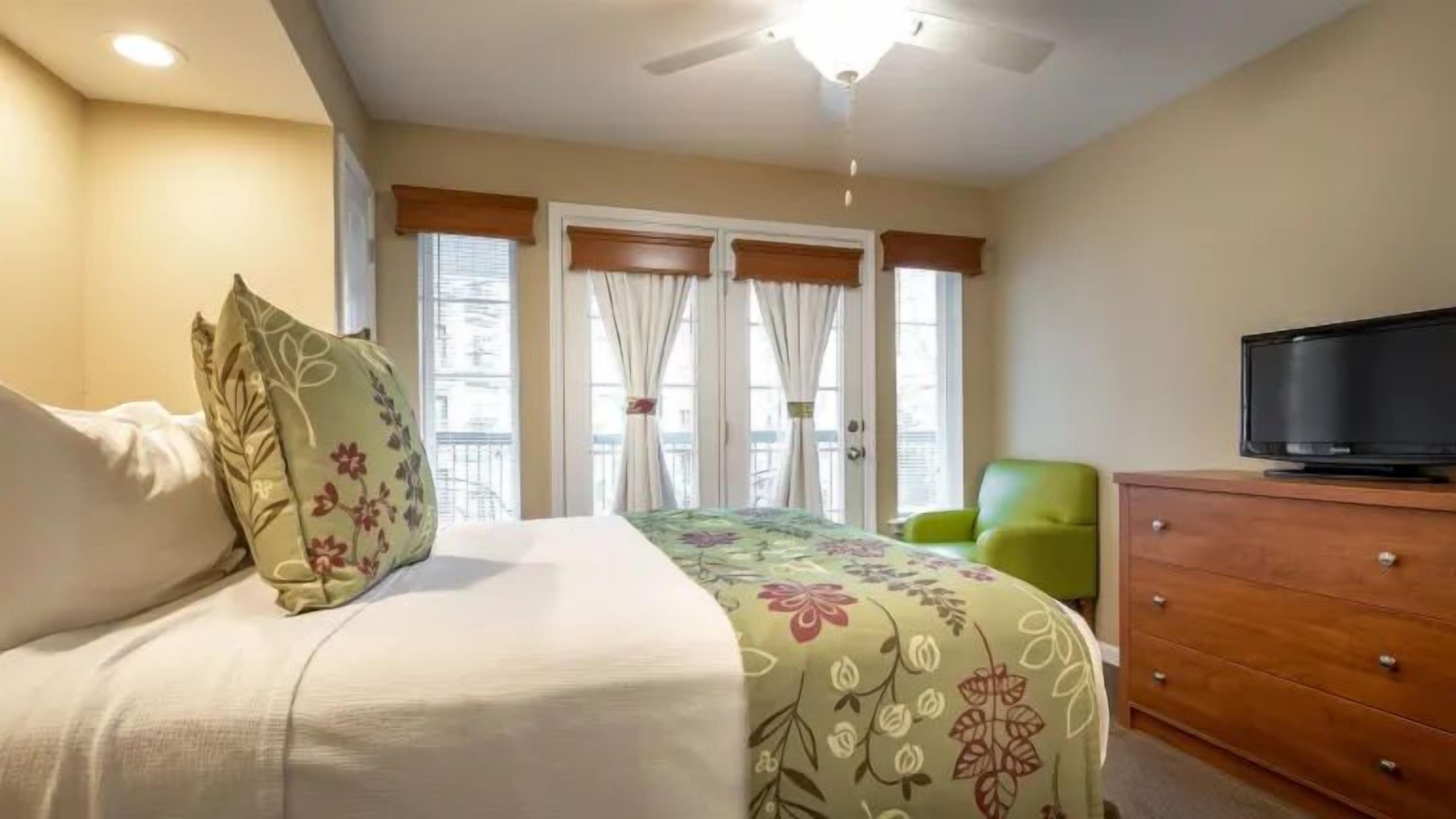 Un dormitorio con una cama, televisión, una silla verde y un aparador de madera.