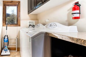 A laundry area with a front-loading washer and dryer, a vacuum cleaner, a fire extinguisher, and a window with a view of trees. - KBM Resorts | In the Heart of Old Town, Walk to Main St., Easy access shuttle to PCMR or DV Resorts, Private Hot Tub! POA-504b (Park City)