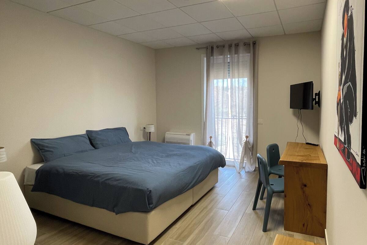 A bedroom with a bed, a desk, a chair, a TV, and a window with curtains.