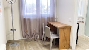 Quarto com escrivaninha de madeira, duas cadeiras, um porta-roupas e janela com cortinas leves.