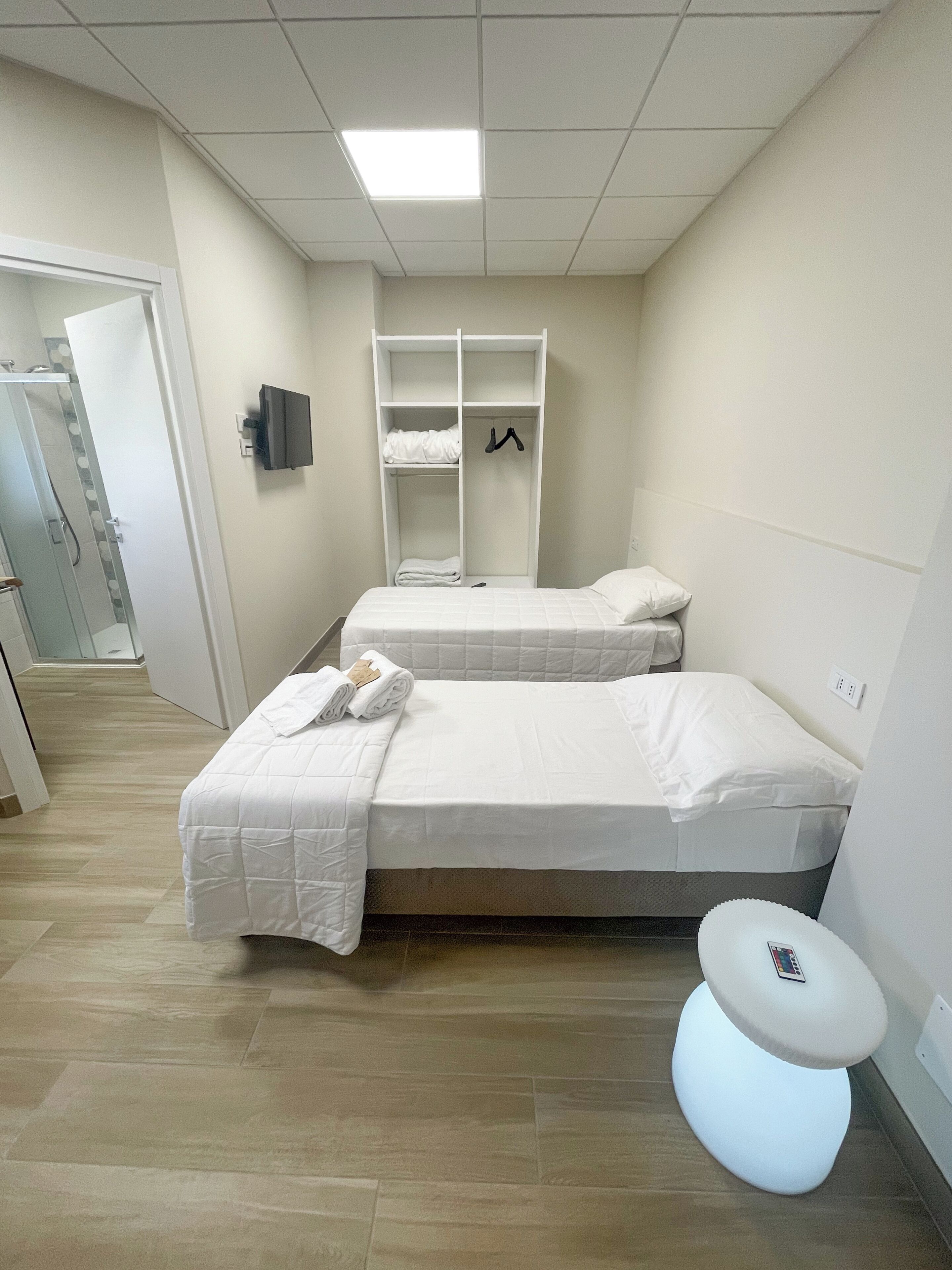 A hotel room with two beds, a wardrobe, a bedside table, and a modern light fixture.