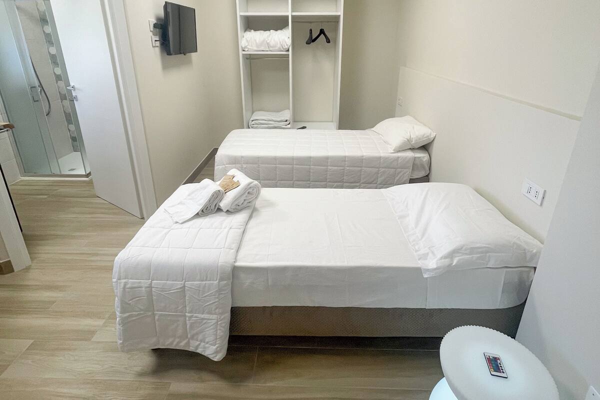 A hotel room with two beds, a wardrobe, a bedside table, and a modern light fixture.