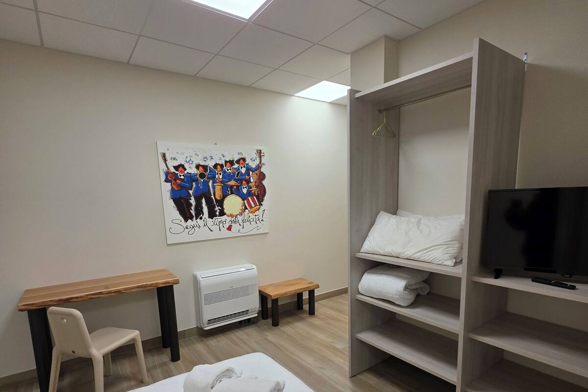A hotel room with a bed, a desk, a chair, a TV, and a shelf with towels.