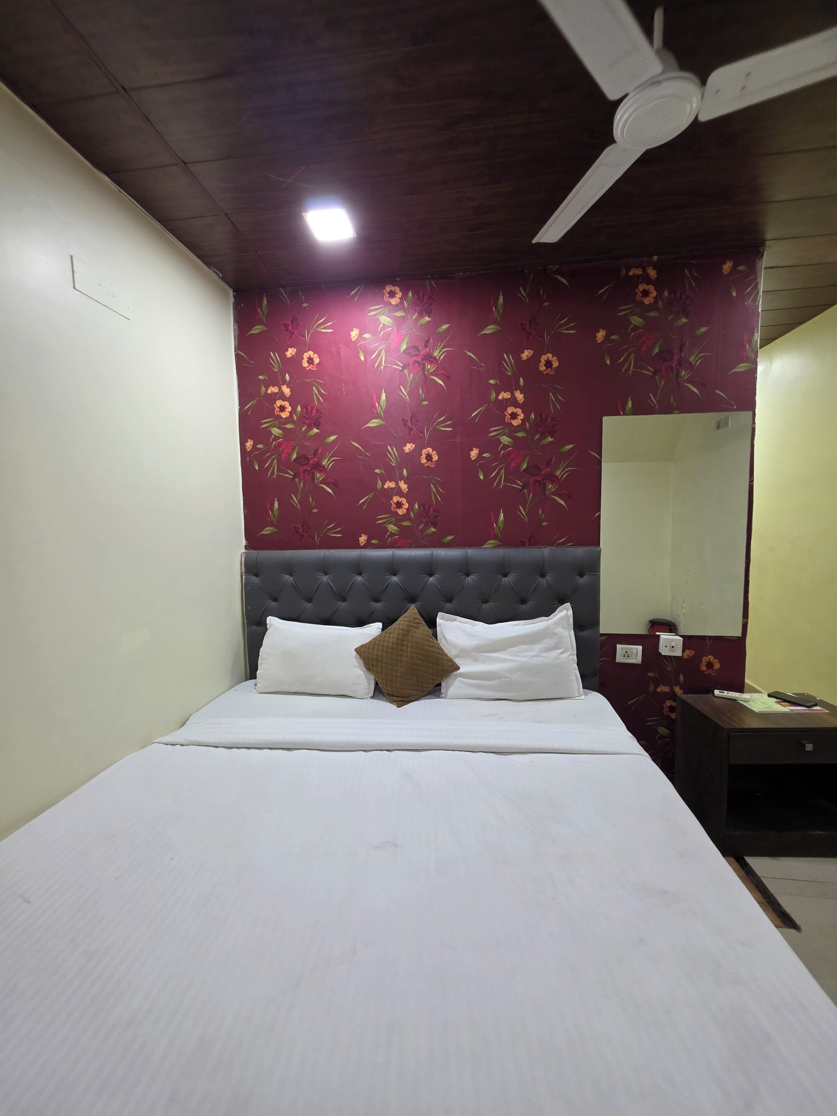 A hotel room with a bed, a ceiling fan, a wall with floral wallpaper, and a mirror.
