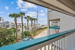 A balcony with a view of a street lined with palm trees and a gas station in the background. - Beachside Escape Steps From Sand Enjoy Ocean Views (Galveston)