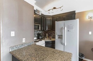 A kitchen with granite countertops, a white refrigerator, dark cabinets, a microwave, and a stove. - Beachside Escape Steps From Sand Enjoy Ocean Views (Galveston)