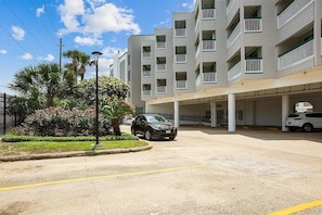 A multi-story building with balconies, a parking area, and a landscaped garden with palm trees and shrubs. - Beachside Escape Steps From Sand Enjoy Ocean Views (Galveston)
