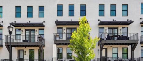 Una hilera de casas urbanas modernas con balcones, un árbol central y cielo despejado.