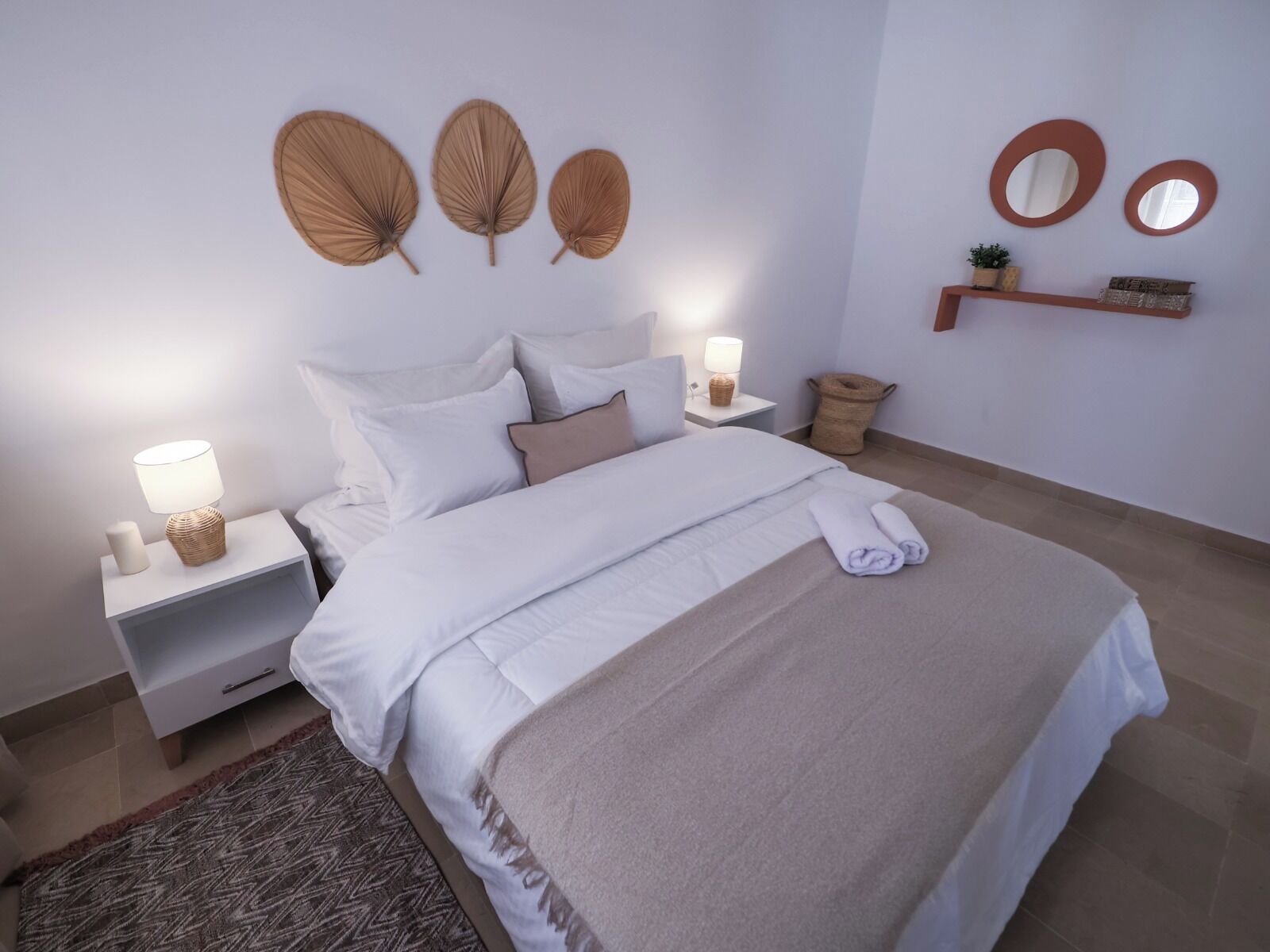 Una cama bien hecha con sábanas blancas, un edredón color beige y una toalla enrollada al pie. Hay dos mesitas de noche con lámparas, una pequeña planta en maceta y un estante montado en la pared con objetos decorativos.