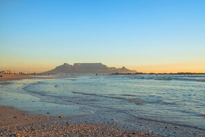 A beach with a large building in the background, calm water, and pebbles on the shore. - Ocean Mist 105 (Cape Town)